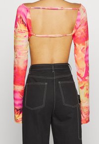 Langärmliges Oberteil mit einem lebhaften pinken und orangen abstrakten Muster, das ein offenes Rückendesign und eine glatte Textur aufweist. Kombiniert mit schwarzen hoch-taillierten Hosen.