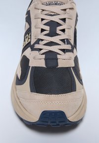 Beige ja tumesinine spordijalats, millel on sametised ja võrgulised materjalid, esile tõstetud paelad ja mustriga tald.