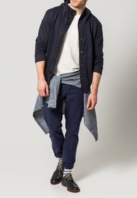 Námořnický mikina s knoflíky a texturovanou kapsou na světle šedém tričku, doplněná tmavě modrými kalhotami a černými botami. Viditelné proužkované ponožky.