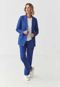 Sinine denimpüüdja kuldsete nööpidega ja sobiva pükstega, millele on kombineeritud valge T-särk ja valged tossud, klassikalise lõike ja laia jalgadega.