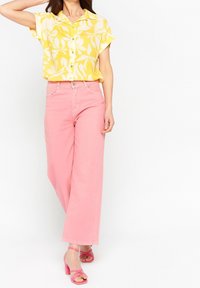 Blusa amarilla de manga corta con patrón floral blanco, combinada con pantalones de pierna ancha rosas y sandalias de tacón rosas.