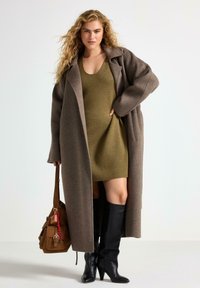 Brun uldfrakke, olivengrøn strikkjole, sorte knæhøje støvler og en brun håndtaske med vedhæng. Modige teksturer og lagdelt look.