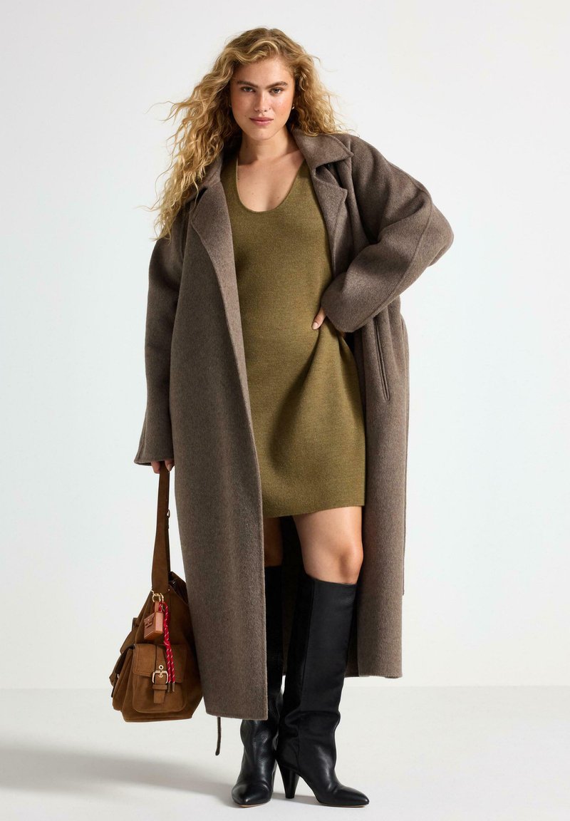 Brun uldfrakke, olivengrøn strikkjole, sorte knæhøje støvler og en brun håndtaske med vedhæng. Modige teksturer og lagdelt look.