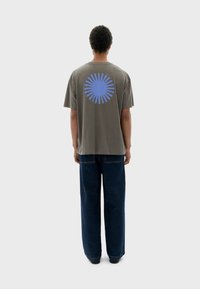 Grauem Baumwoll-T-Shirt mit einem blauen Sonnenstrahl-Design auf dem Rücken. Kombiniert mit dunkelblauen weiten Jeans, von hinten gezeigt.