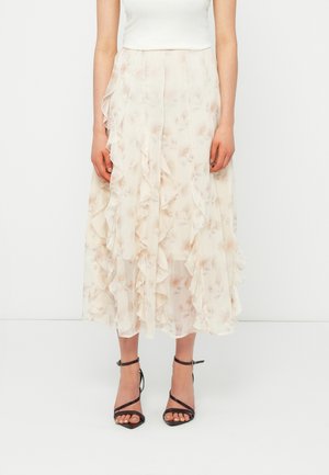 Naine kannab keskmise pikkusega kreemika lillemustriga volditud seelikut, valget toppi ja musti rihmikandvaid kõrge kontsaga sandaale, seistes valge tausta ees.