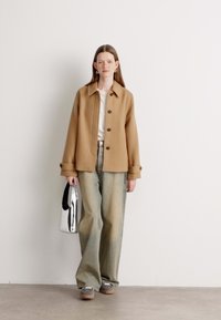Brun uldjakke med store knapper, beige top, brede blå jeans og sølvfarvet totebag. Model står på en neutral baggrund.