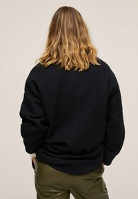 Musta, teksturoidusta kankaasta valmistettu rento neule, jossa on leveät hihat, näkymä takaa. Yhdistetty istuviin oliivinvihreisiin housuihin.