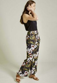 Pantalons larges floraux en tissu noir, ornés de fleurs blanches et orange ainsi que de feuilles vertes. Associés à un débardeur noir moulant et des sandales marron.