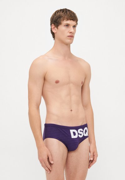 Jeune homme aux cheveux bruns courts portant un slip de bain violet avec un grand logo blanc "DSQ" sur fond blanc.