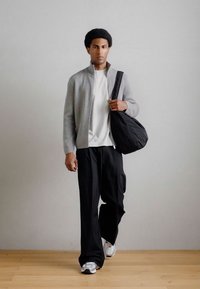 Ανοιχτό γκρι φούτερ με φερμουάρ πάνω από ένα λευκό πουκάμισο, συνδυασμένο με μαύρο παντελόνι σε φαρδιά γραμμή και γκρι αθλητικά παπούτσια. Μια μαύρη τσάντα είναι κρεμασμένη στον έναν ώμο.