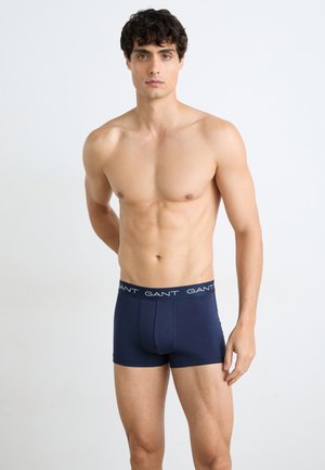 Hemdloser Mann steht vor schlichten Hintergrund und trägt marineblaue Boxer mit GANT-Logo am Bund.