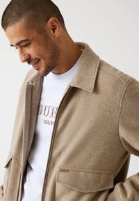 Lichtbruine wollen blend jas met een kraag, ritsluiting en twee zakken aan de voorkant. Gedragen over een wit t-shirt met subtiele blindgeperste branding.