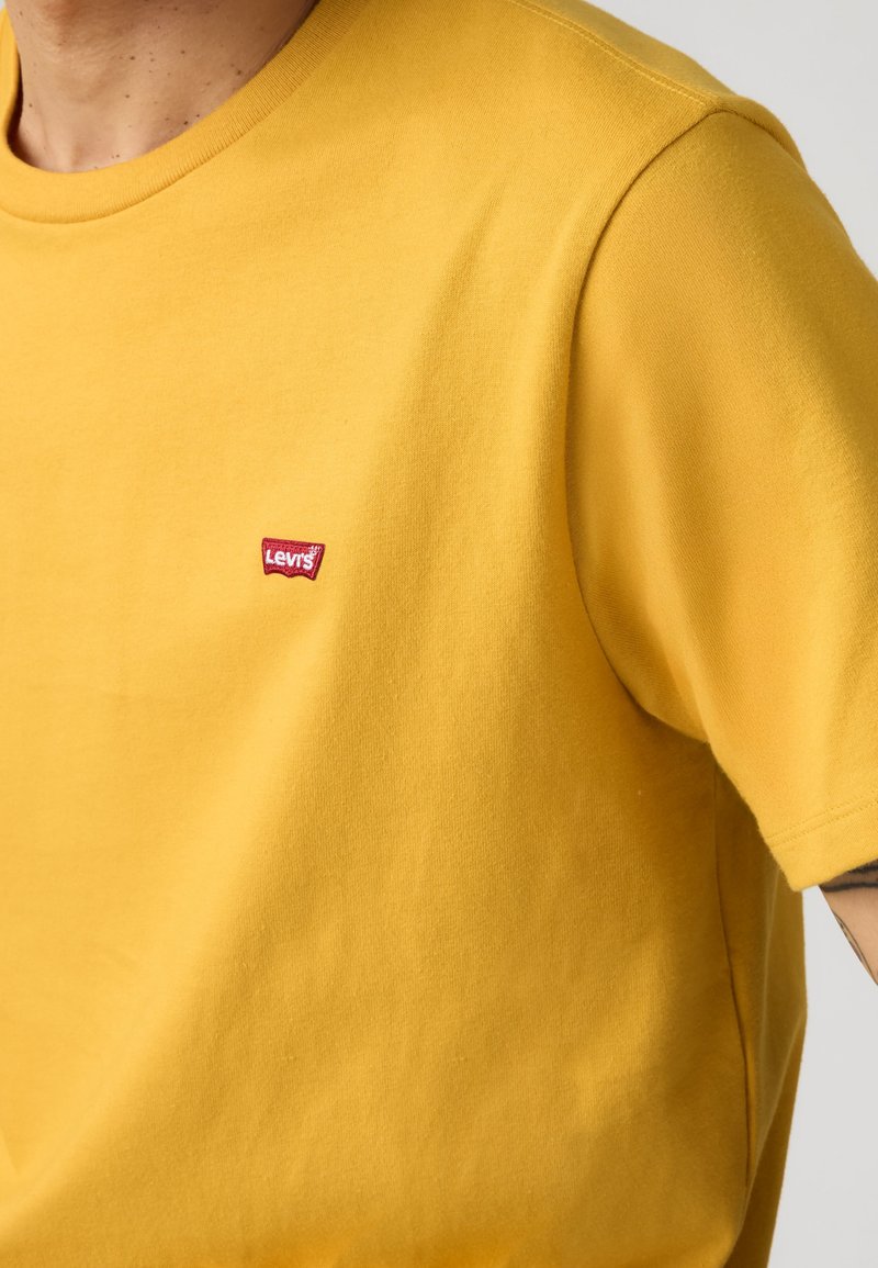 T-shirt jaune à manches courtes avec un petit logo rouge Levi's sur la poitrine gauche, porté par une personne avec un tatouage partiel visible sur le bras.