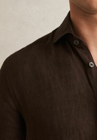 Camisa de lino marrón con cuello abotonado y dos botones visibles, que presenta una tela ligeramente texturizada y un corte relajado.
