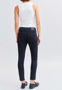 Žena v bílém bezrukávovém topu, černých slim-fit džínách a černých loaferkách, stojící s obratem zády k fotoaparátu.