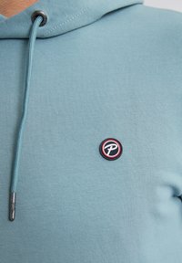 Sudadera azul claro hecha de una mezcla de algodón, con un parche de logo redondo en rojo, negro y blanco en la zona del pecho izquierdo, y con detalle de cordón.