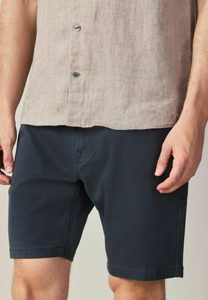 Muž oblečený v béžovej košeli na gombíky zastrčenej do čiernych krátkych nohavíc po kolená, stojaci s rukami voľne pri tele.