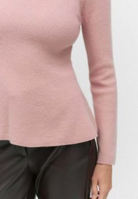 Pull en maille rose à côtes avec des manches longues, présentant un ourlet peplum évasé et des détails de couture subtils sur les côtés. Associé à un pantalon sombre.
