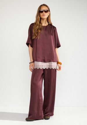 Femme portant un haut en satin bordeaux ample et un pantalon à larges jambes, de grandes lunettes de soleil et des bracelets, debout devant un fond clair uni.