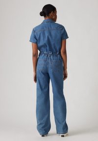 Blå denim jumpsuit med korte ærmer, elastisk talje og vide bukser. Har baglommer og kontrasterende sømdetaljer.