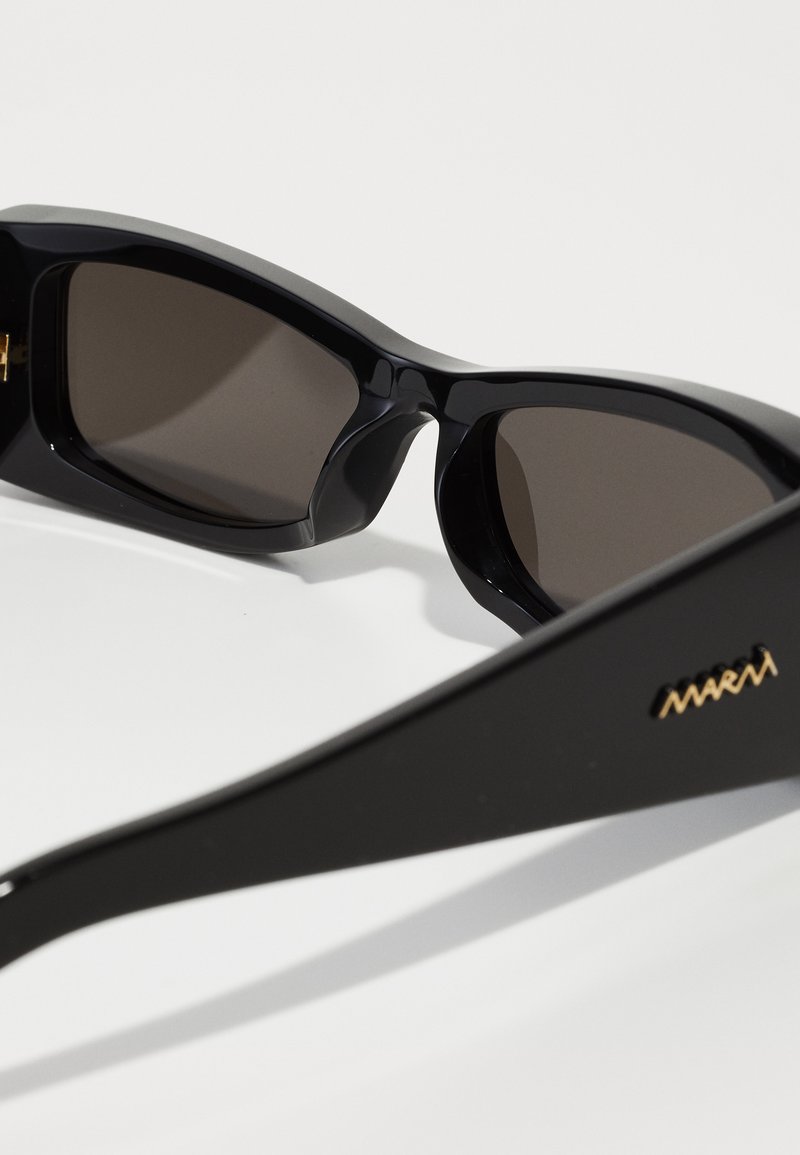 Lunettes de soleil noires avec une monture en plastique lisse, des verres foncés et un détail en logo doré sur la branche. Design angulaire avec des côtés larges.