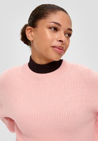 Pull en tricot rose clair avec une finition texturée, présentant un col roulé côtelé marron foncé et une coupe décontractée.