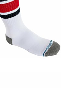 Weißer Socken mit roten und schwarzen Streifen oben, grauer Ferse und Zehen. Besitzt einen strukturierten Stoff und Branding am Bündchen.