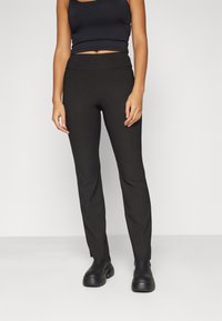 Pantalons noirs ajustés en matériau doux et extensible ; taille haute avec une bande lisse et plate ; design de jambe fuselée. Associés à des bottes noires à la cheville.