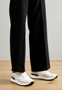 Sportowe sneakersy mają białą cholewkę z szarymi zamszowymi akcentami, czarną podeszwę i sznurowany design. Noszone z luźnymi czarnymi spodniami na drewnianej podłodze.