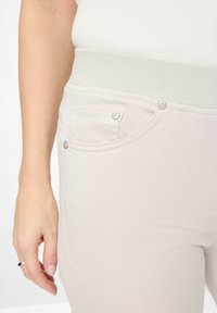 Pantalon beige clair avec une taille élastique, deux poches avant avec boutons-pression, et une texture en coton lisse.