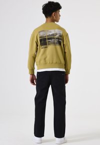 Sudadera verde oliva con un estampado gráfico en la parte posterior, presenta puños y dobladillo de canalé. Combinada con pantalones negros y zapatillas blancas.