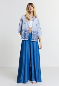 Frau in einem blauen bestickten Hemd mit Knöpfen über einem weißen Crop-Top und weit geschnittenen blauen Hosen mit weißen offenen Sandalen, stehend vor einem weißen Hintergrund.