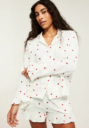 Femme portant un ensemble de pyjama blanc à boutons avec des motifs de cœurs rouges, comprenant des manches longues et un short froncé.