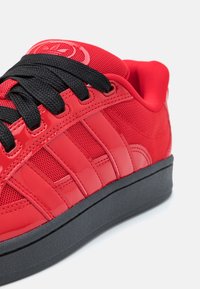 Zapatilla deportiva roja con una combinación de materiales brillantes y de malla, cordones negros y suela de goma. Destaca el diseño de tres rayas en un panel elevado.