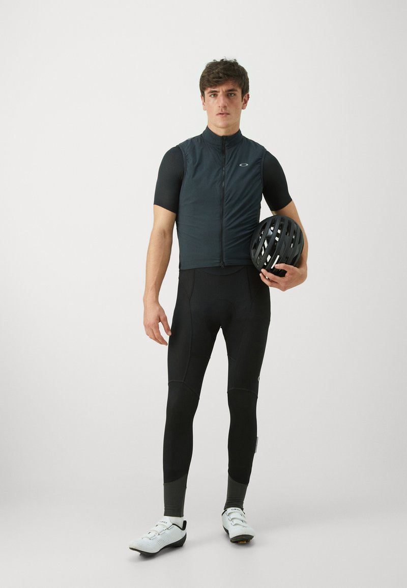 Schwarze Fahrradweste mit kurzen Ärmeln, enganliegende schwarze Radlerhosen und weiße Fahrradschuhe. Das Modell hält einen schwarzen Helm und steht vor einem neutralen Hintergrund.