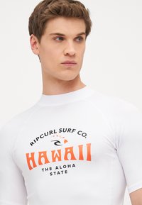 Hvit kortermet rashguard med et dristig svart og oransje design som viser teksten "HAWAII" med teksten "RIP CURL SURF CO." over.