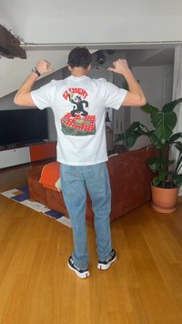 Άνδρας που στέκεται μέσα σε κλειστό χώρο, φοράει λευκό μπλουζάκι με γραφικά, με καρτούν και το κείμενο "Element", μπλε τζιν και μαύρα και λευκά αθλητικά παπούτσια.