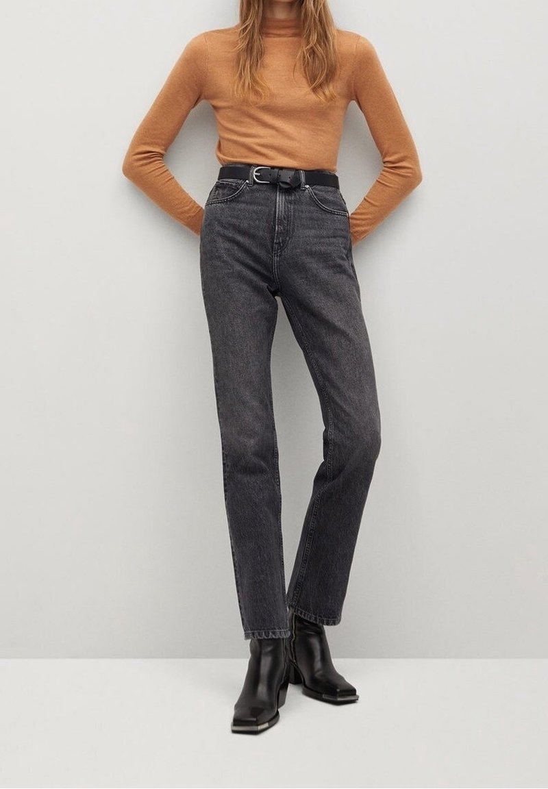 Femme portant un haut marron à manches longues, un jean noir taille haute avec ceinture, et des bottines noires, debout contre un mur clair uni.