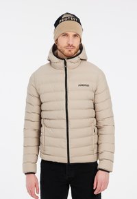 Veste doudoune beige avec des coutures horizontales, fermeture éclair et capuche. Comprend des accents noirs et un logo. Associée à un bonnet en tricot beige et bleu marine.