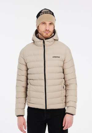 Veste doudoune beige avec des coutures horizontales, fermeture éclair et capuche. Comprend des accents noirs et un logo. Associée à un bonnet en tricot beige et bleu marine.