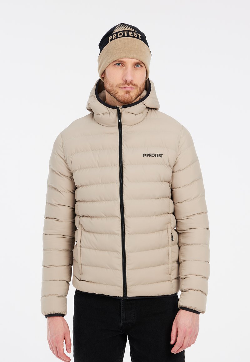 Veste doudoune beige avec des coutures horizontales, fermeture éclair et capuche. Comprend des accents noirs et un logo. Associée à un bonnet en tricot beige et bleu marine.