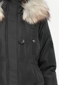 Črna parka s kapuco obloženo s krznom, dvojno gumbi, elastičnim pasom in teksturirano tkanino. Vključuje logotip na rokavu.