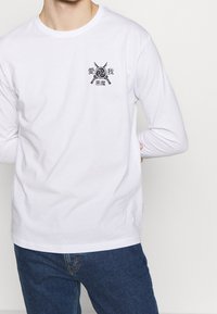 Weißes Langarmhemd mit kleinem schwarzem japanischen Design von gekreuzten Schwertern und rundem Emblem auf der linken Brust, getragen von einer Person in blauen Jeans.