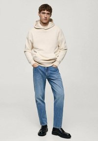 Beige hoodie met een kangoeroezak, gecombineerd met lichtblauwe jeans en zwarte boots. Model staat rechtop, handen in de zakken, neutrale achtergrond.