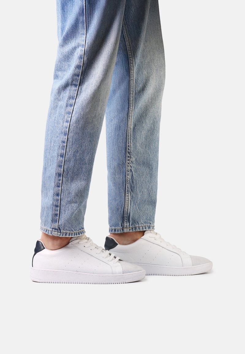 Jambes portant un jean bleu clair et des baskets basses blanches avec un détail noir au talon, debout sur un fond blanc uni.