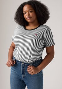 Kvinne med krøllete hår iført en svart-hvitt stripete Levi's t-skjorte og blå jeans, som holder i midjen, stående mot en lys bakgrunn.