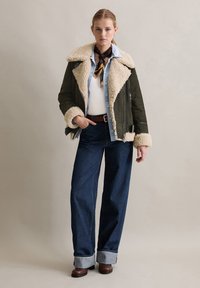Olijfgroene shearling-voering jack met een lichtblauw spijkerhemd, een crèmekleurige trui, donkerblauwe wijde jeans en bruine enkellaarsjes.