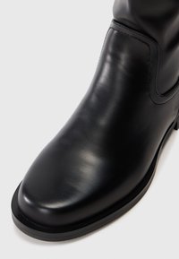 Bota de tornozelo em couro preto com bico arredondado e costura minimalista em fundo branco.