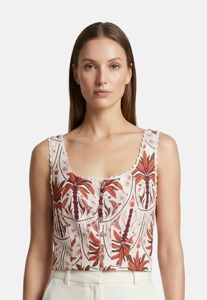 Woman with straight brown hair wears sleeveless white crop top with red and brown leaf pattern and button details, paired with white pants.