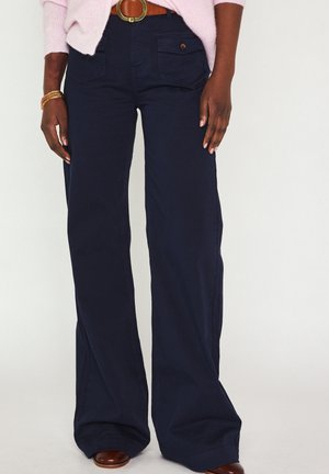 Personne portant un pantalon large bleu marine foncé, une ceinture en cuir marron, un pull rose et des chaussures marron, debout avec les mains détendues sur les côtés.